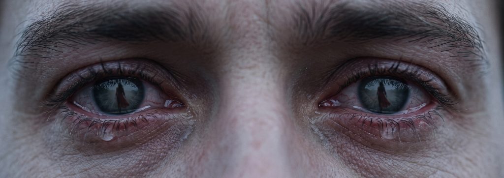 Nahaufnahme der tränenerfüllten Augen eines Mannes, in deren Spiegelung die Silhouette einer weggehenden Frau zu erkennen ist.